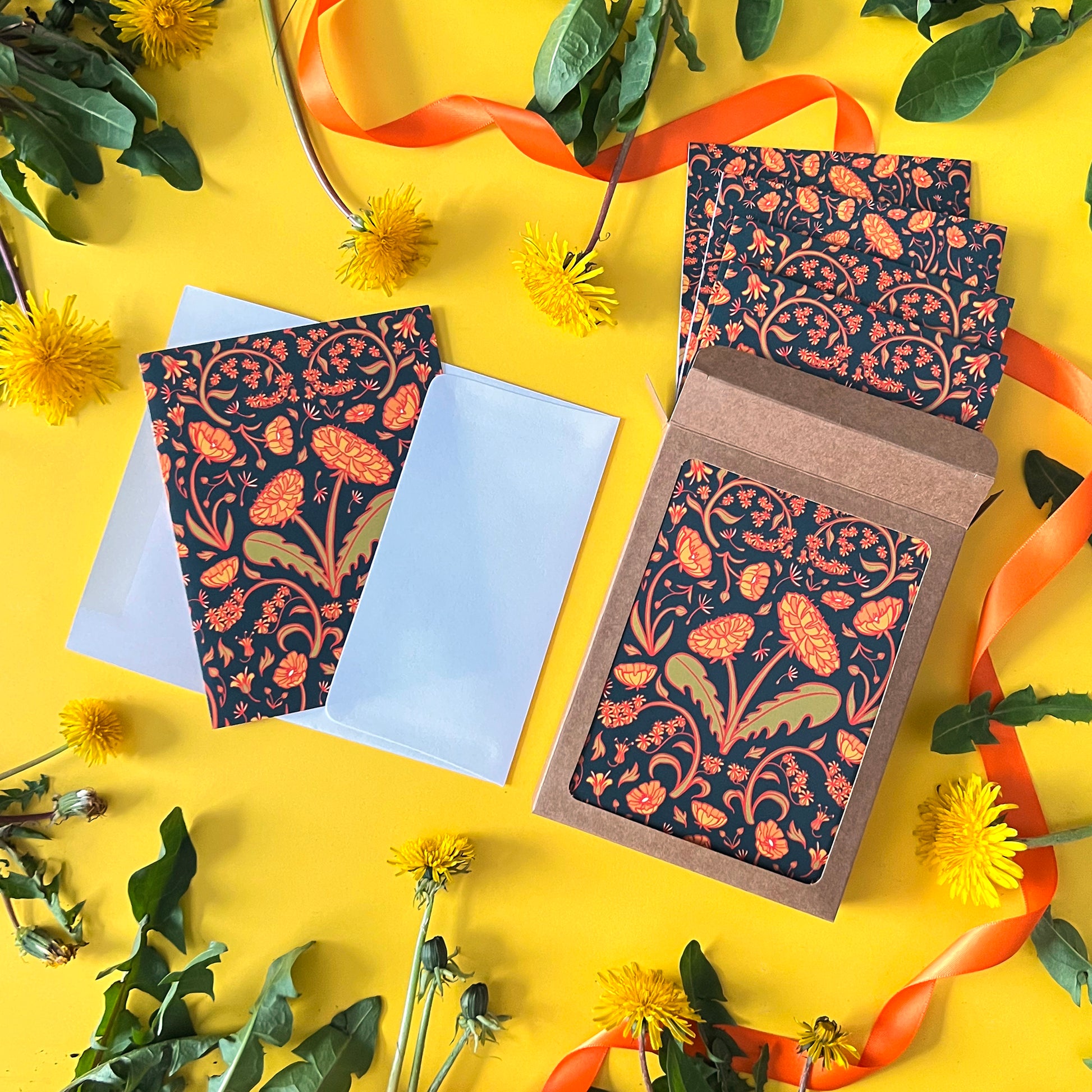 A box of dandelion patterned note cards is next to the same card peeking out ofan envelope. They are on yellow woth dandelion leaves, flowers, and orange ribbon.