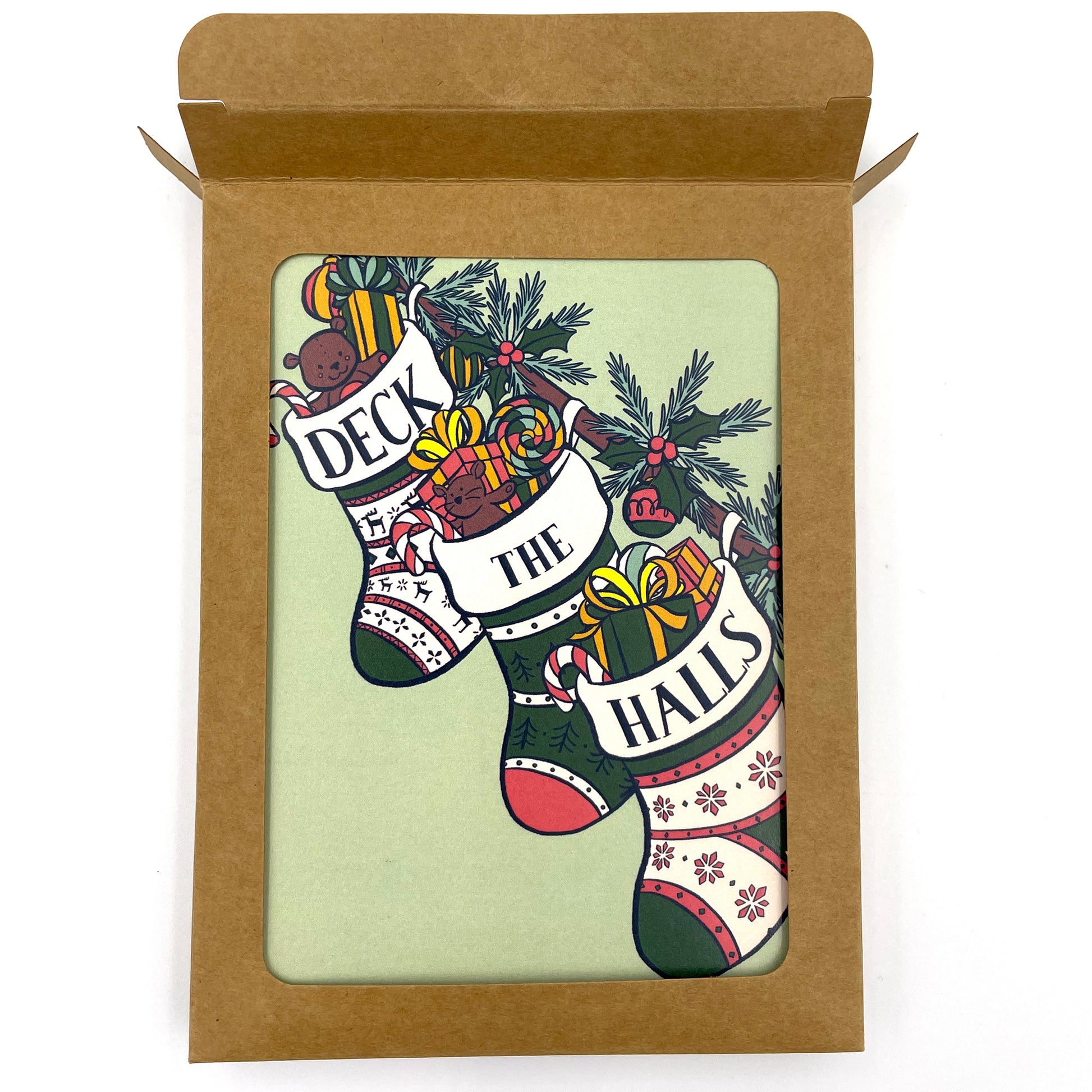 A mint green card shows three stockings stuffed with toys, gifts, and candy along a banister. The words Deck the Halls are split among the tops of each stocking. The card shows through a window in a kraft paper box and the top of the box is open.