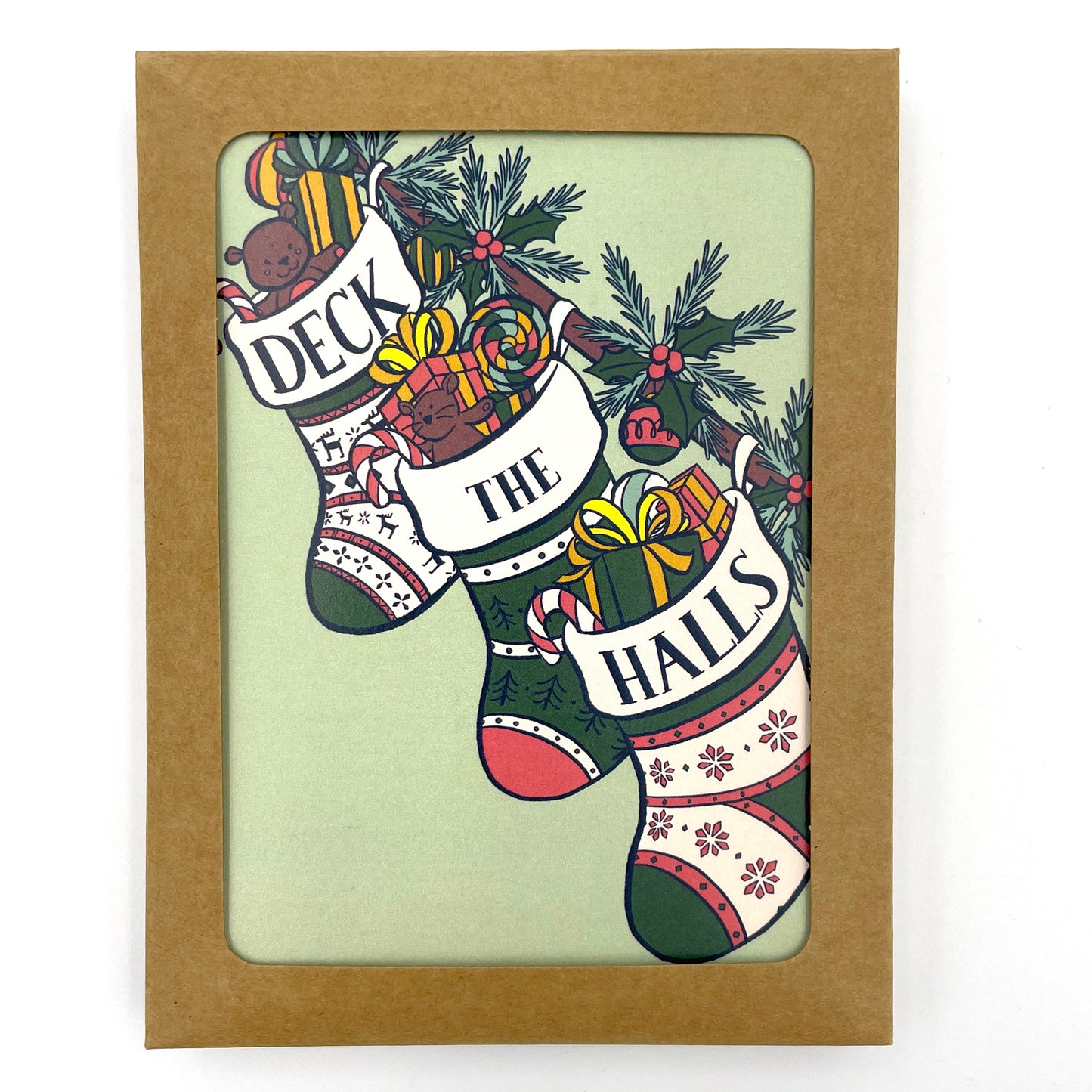 A mint green card shows three stockings stuffed with toys, gifts, and candy along a banister. The words Deck the Halls are split among the tops of each stocking. The card shows through a window in a kraft paper box.