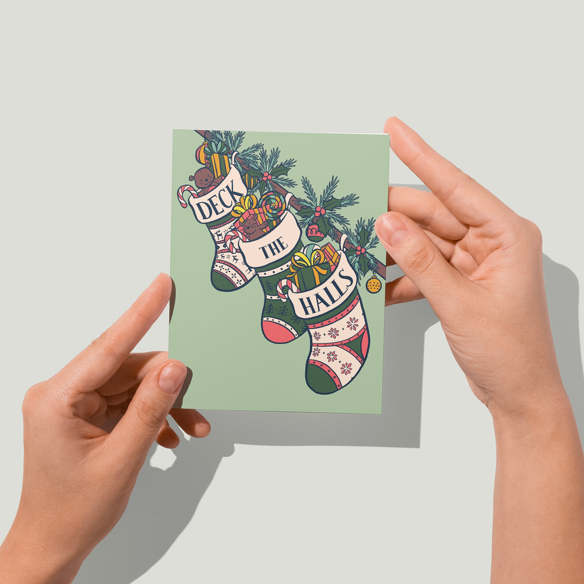 A mint green card shows three stockings stuffed with toys, gifts, and candy along a banister. The words Deck the Halls are split among the tops of each stocking. A pair of hands holds the card up in front of the camera.