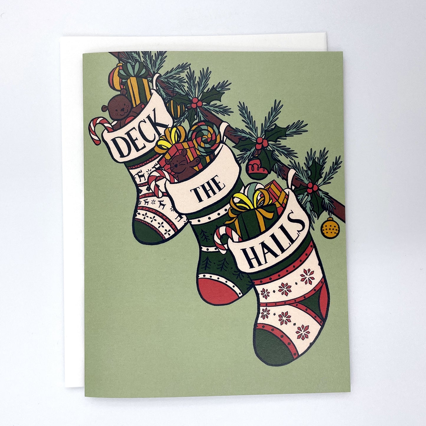 A mint green card shows three stockings stuffed with toys, gifts, and candy along a banister. The words Deck the Halls are split among the tops of each stocking. The card is against a white envelope on a white background