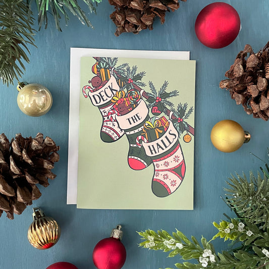 A mint green card shows three stockings stuffed with toys, gifts, and candy along a banister. The words Deck the Halls are split among the tops of each stocking. The card is surrounded by faux greenery, Christmas ornaments, and pinecones.