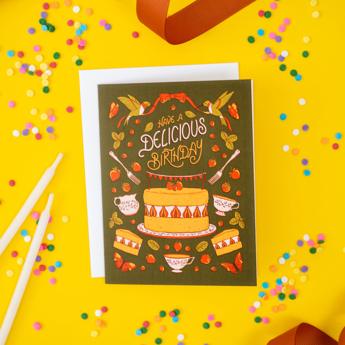 A green greeting card shows birds, butterflies, teacups, and the words Have a Delicious Birthday in hand-lettering. The card is on a yellow  background strewn with confetti.