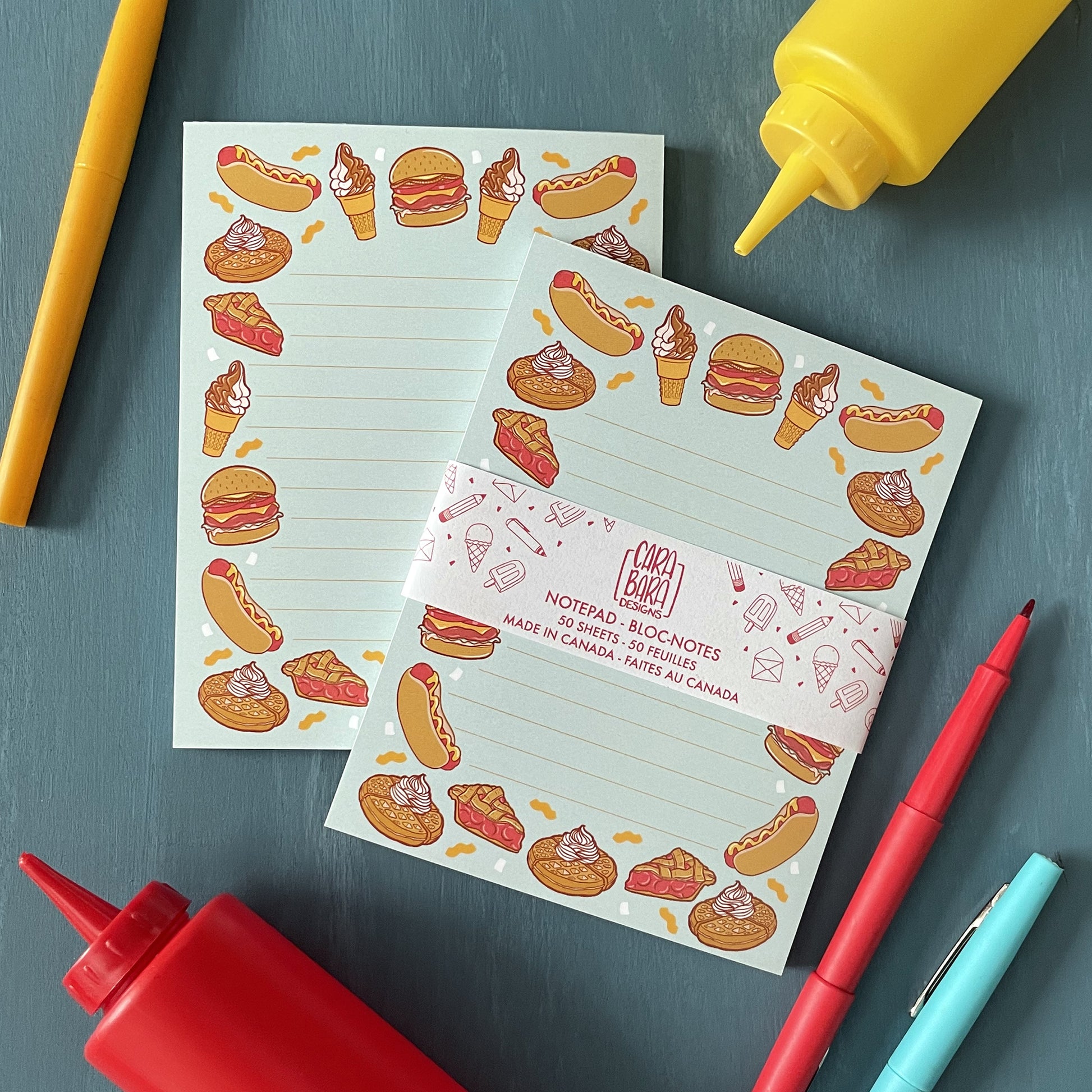 A pair of blue lined notepads feature illustrations of burgers, hot dogs, ice cream, waffles, and pie. The 50-page notepad is on a blue surface with ketchup and mustard squeeze bottles and red, yellow and blue pens. The front notepad is packaged in a belly band with the Carabara Designs logo, indicating the notepad is 50 pages and made in Canada.