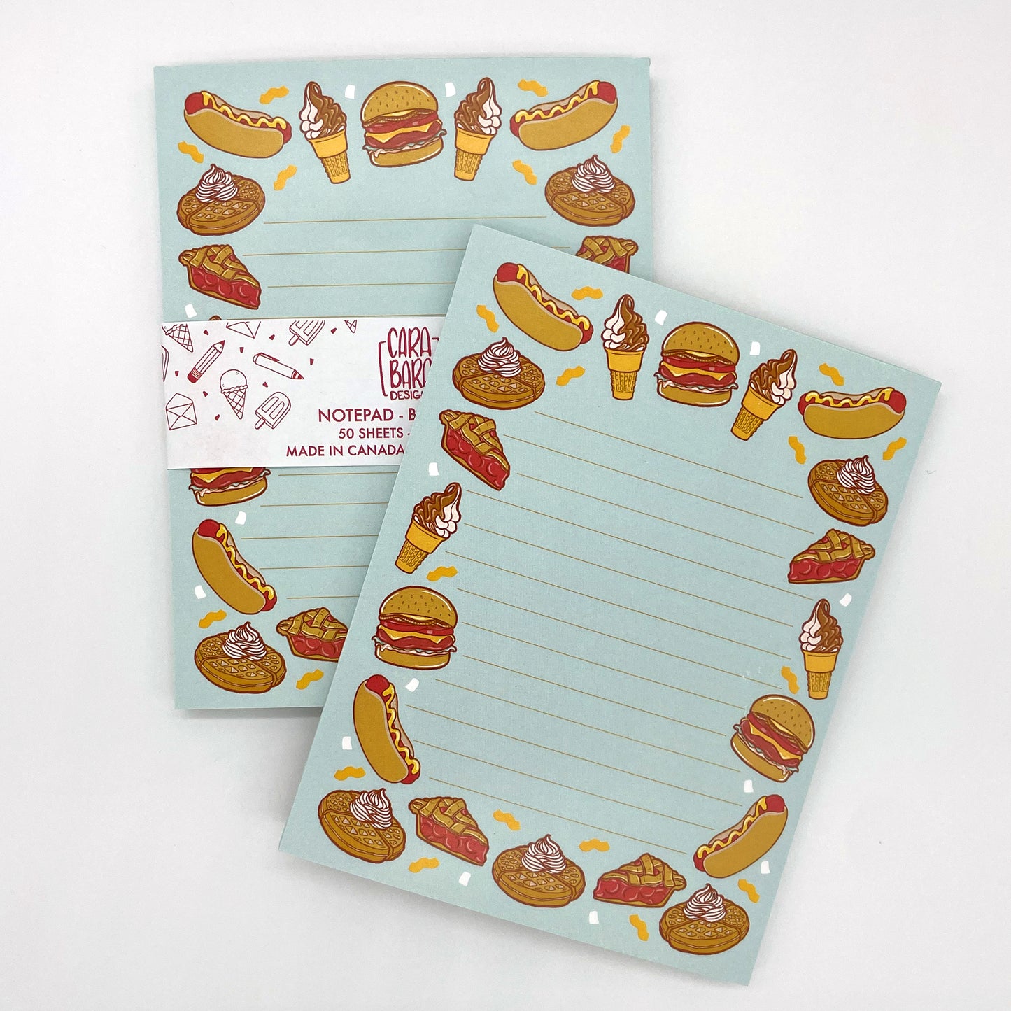 A pair of blue lined notepads feature illustrations of burgers, hot dogs, waffles, ice cream and pie. The back notepad is packaged in a belly band with the Carabara Designs logo, indicating the notepad is 50 pages and made in Canada.