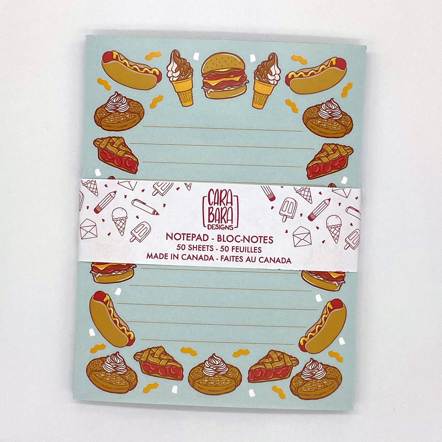 A blue lined notepad features illustrations of burgers, hot dogs, waffles, ice cream and pie. The notepad is packaged in a belly band featuring the Carabara Designs logo and indicating the notepad is 50 pages and made in Canada.