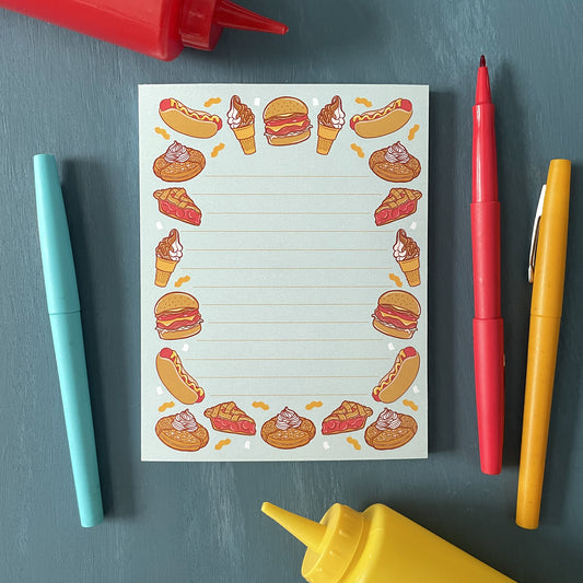 A blue lined notepad features illustrations of burgers, hot dogs, ice cream, waffles, and pie. The 50-page notepad is on a blue surface with ketchup and mustard squeeze bottles and red, yellow and blue pens.