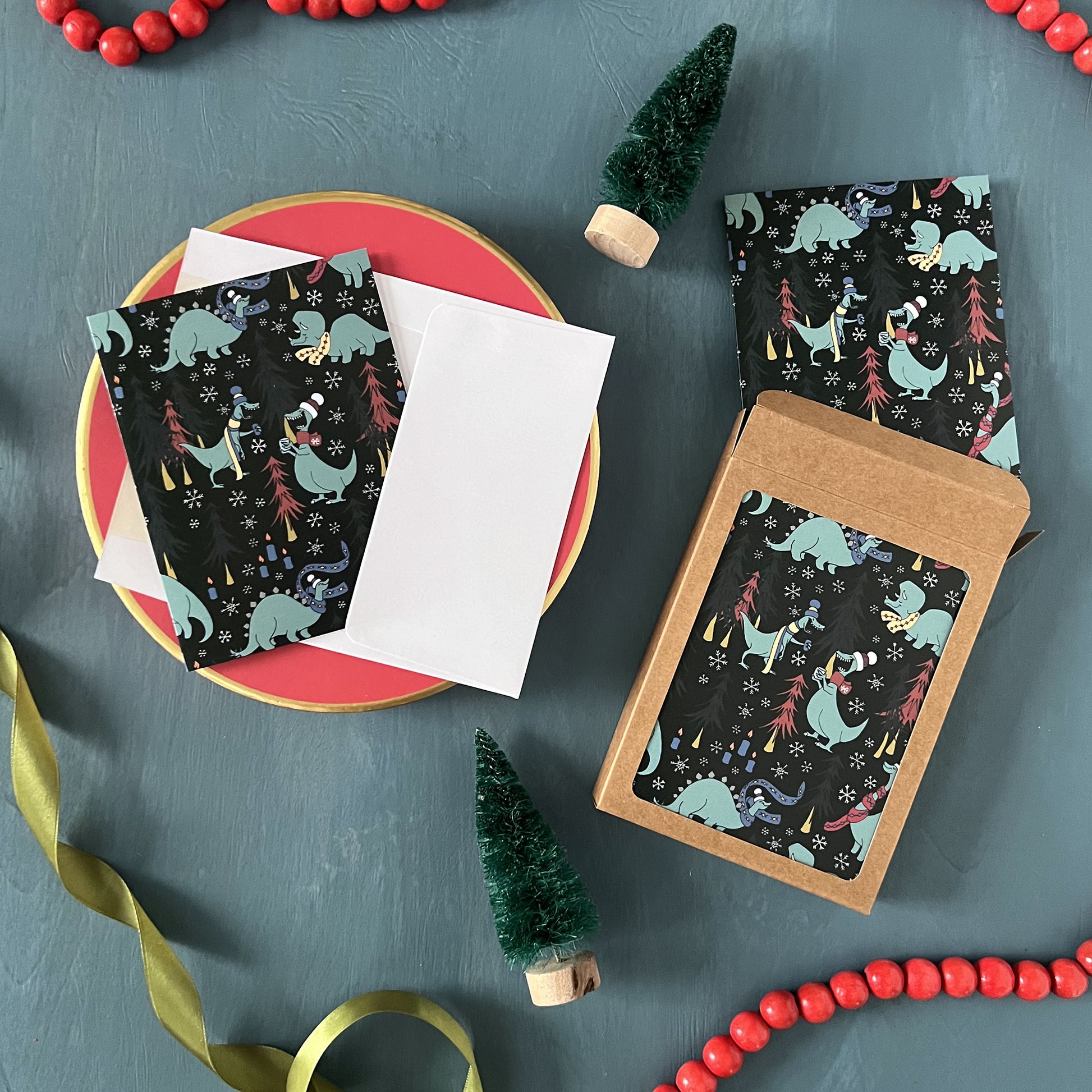 Christmas greeting cards patterned with cozy dinosaurs in the woods appear on a festive blue background with red beads, green ribbon, and bottle brush trees.
