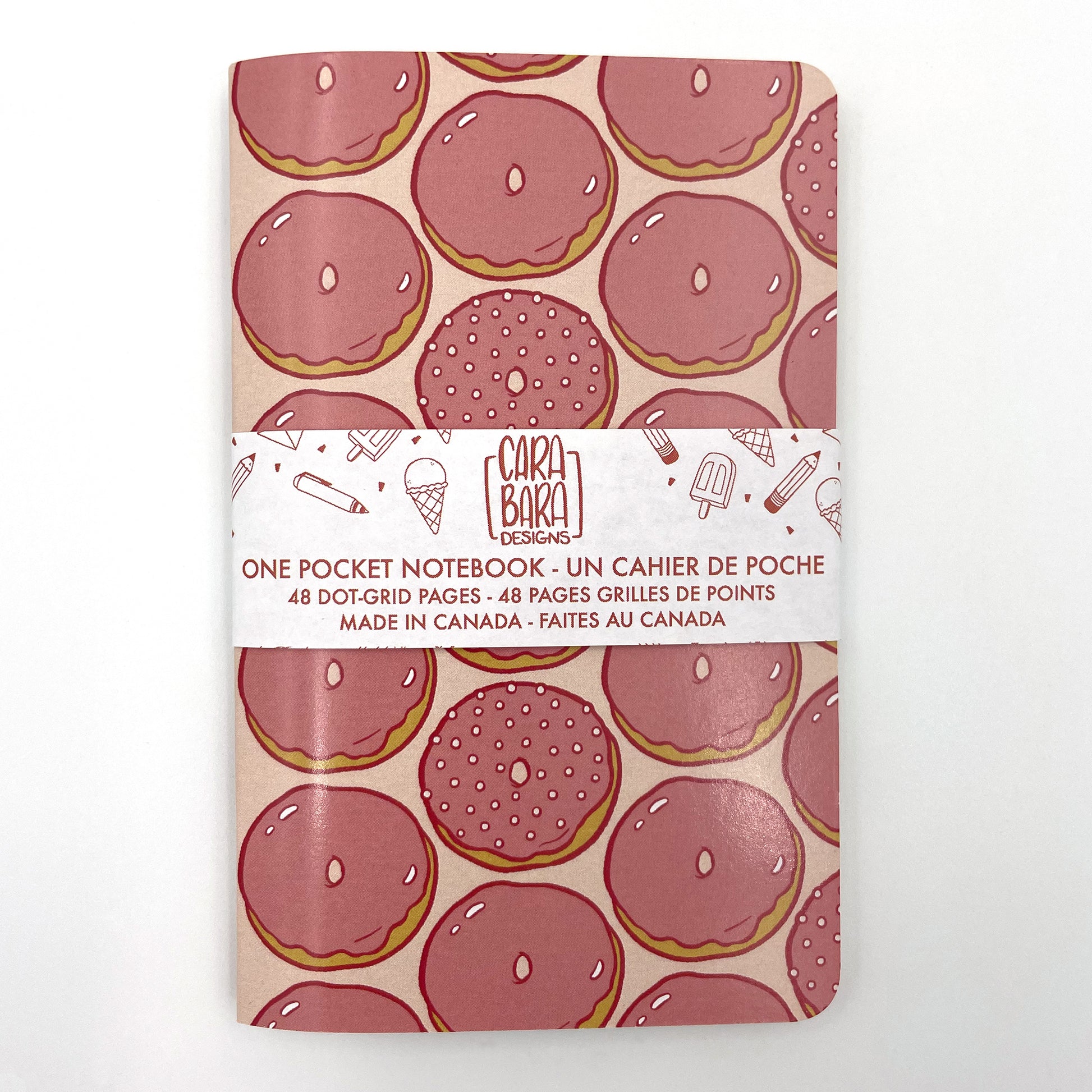 A small closed notebook has a cover patterned with pink glazed doughnuts aligned in vertical stripes. The notebook is bound with a Carabara Designs belly band indicating in both French and English that it is a pocket notebook containing 48 dot grid pages and is made in Canada.