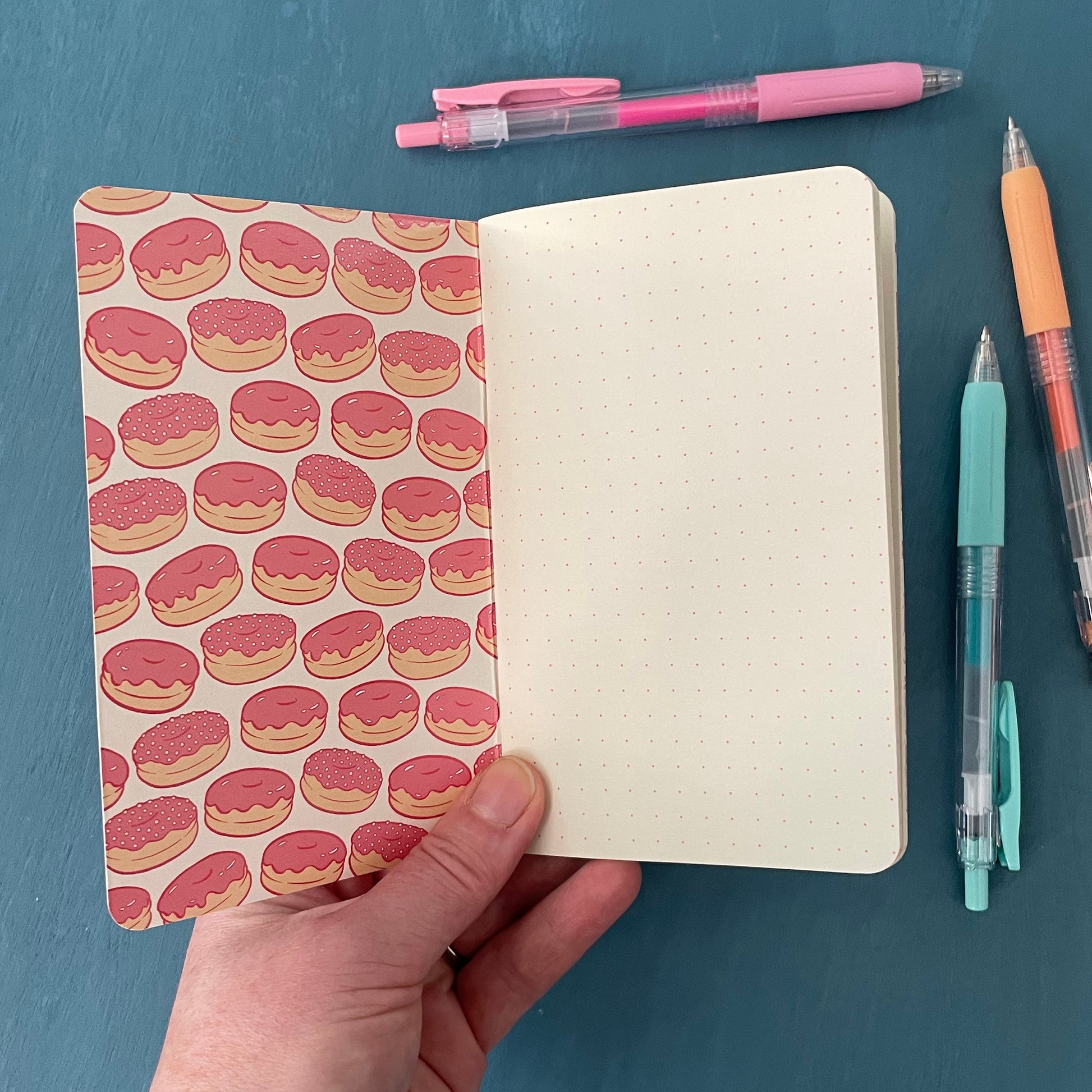 A pocket-sized notebook is open to its first page. The inside cover is patterned with profiles of pink glazed doughnuts and its first page is covered with a deep raspberry dot grid on creamy paper stock. The notebook is surrounded by pens.