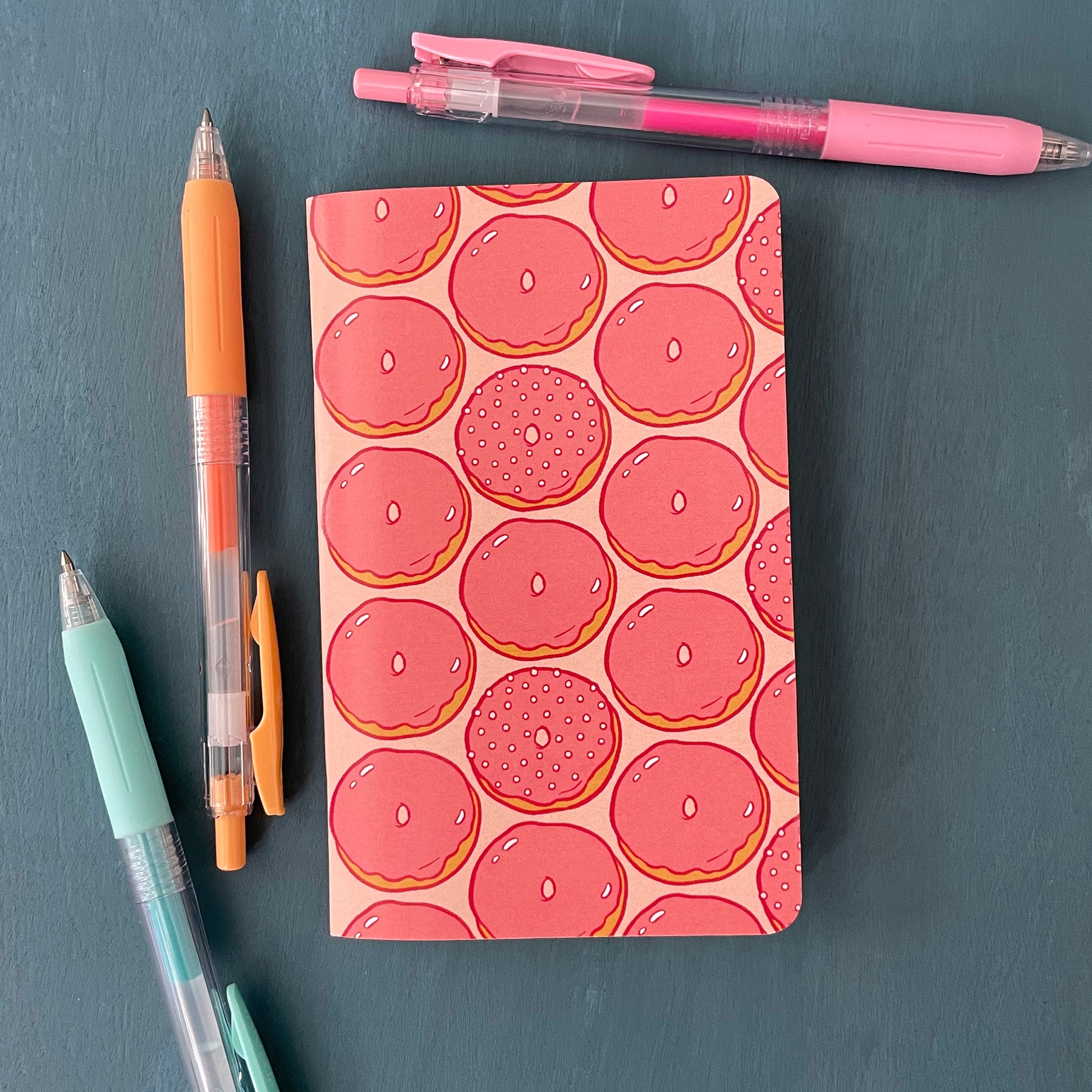 A pocket-sized notebook has a pink cover patterned with pink glazed doughnuts. It sits on a blue background surrounded by pens.