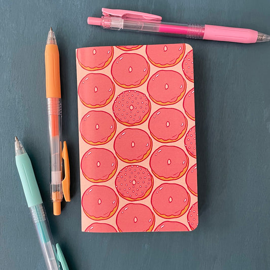 A pocket-sized notebook has a pink cover patterned with pink glazed doughnuts. It sits on a blue background surrounded by pens.