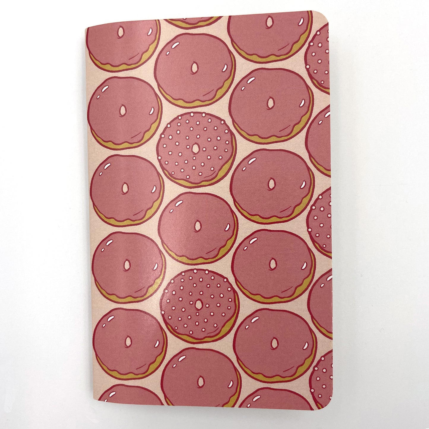 A small closed notebook has a cover patterned with pink glazed doughnuts aligned in vertical stripes.