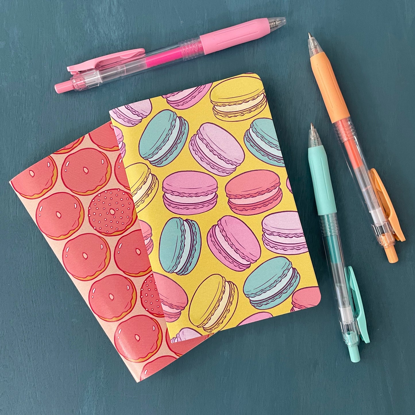 A pocket-sized notebook has a yellow cover patterned with French macarons and sits atop another pocket notebook patterned with pink glazed doughnuts. It sits on a blue background surrounded by pens.