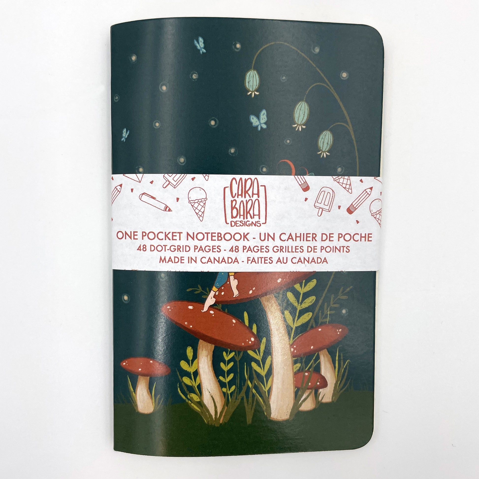 A small closed notebook has a cover that shows a fairy sitting on a mushroom in a field of other mushrooms, with butterflies and dots in a dark sky. The notebook is bound with a Carabara Designs belly band indicating in both French and English that it is a pocket notebook containing 48 dot grid pages and is made in Canada.