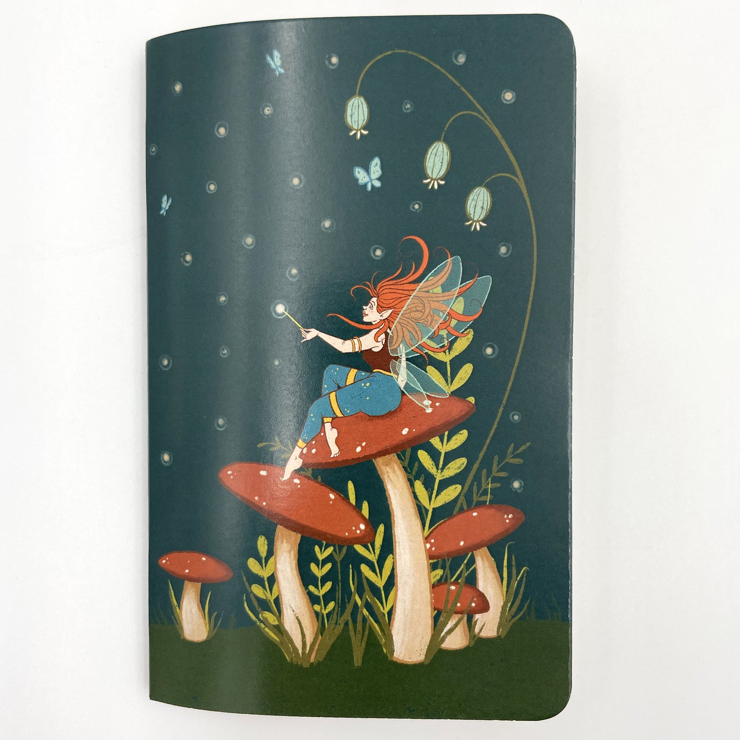 A small closed notebook has a cover that shows a fairy sitting on a mushroom in a field of other mushrooms, with butterflies and dots in a dark sky.