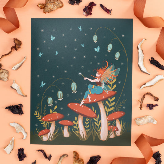 An art print of a fairy on mushrooms at night is on orange with dried wild mushrooms and brown ribbon.