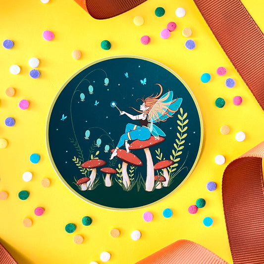 A round sticker features a fairy with a wand sitting on top of mushrooms. It is on yellow with ribbon and confetti.