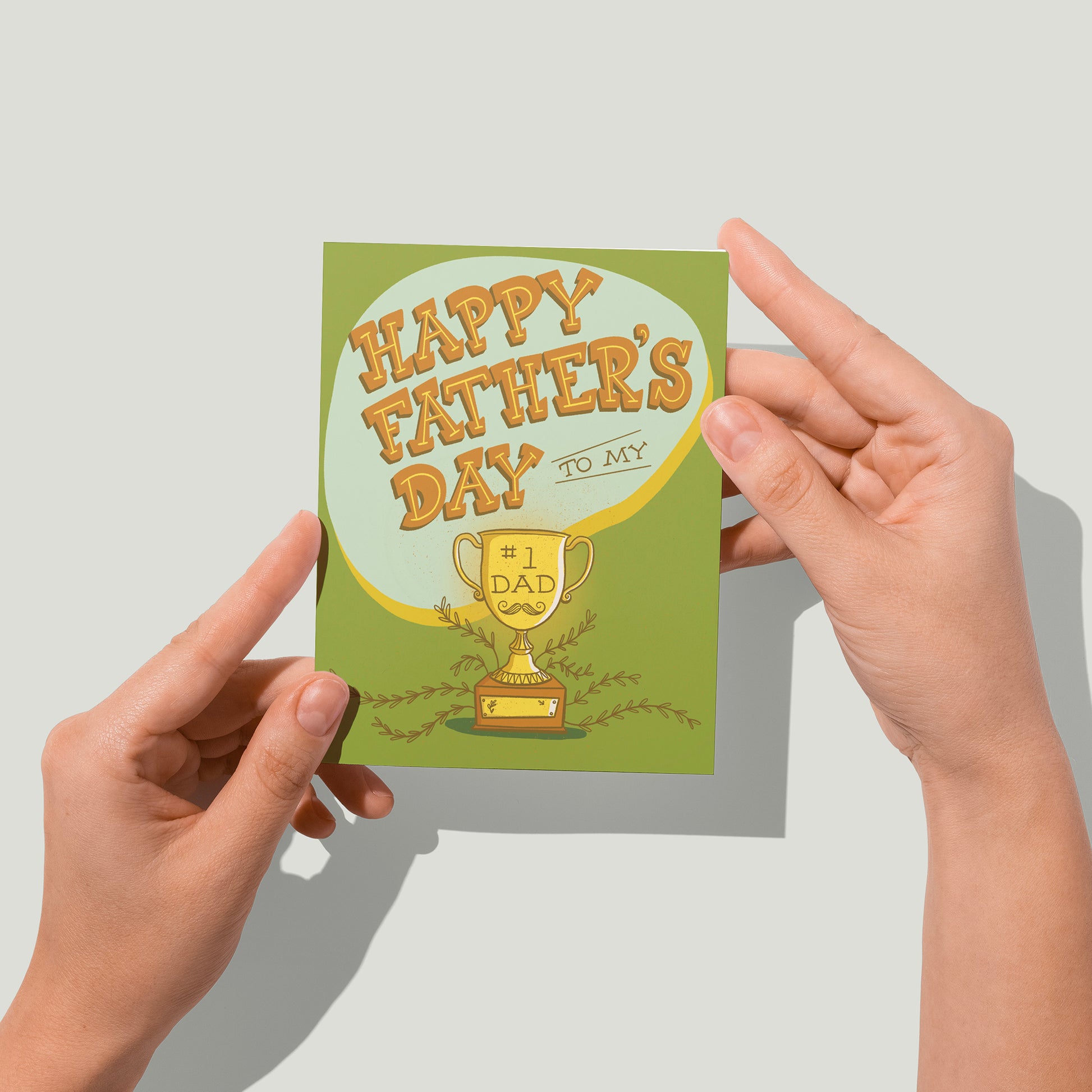 A green greeting card shows hand-lettered text reading Happy Father's Day to my in midcentury-inspired hand-lettering on a blue blob. Below the letters is a shining golden trophy on which it says Number One Dad. The trophy also shows an image of a mustache and is surrounded by vines. A blank plaque is on its base. A pair of hands holds the card up in front of the camera.