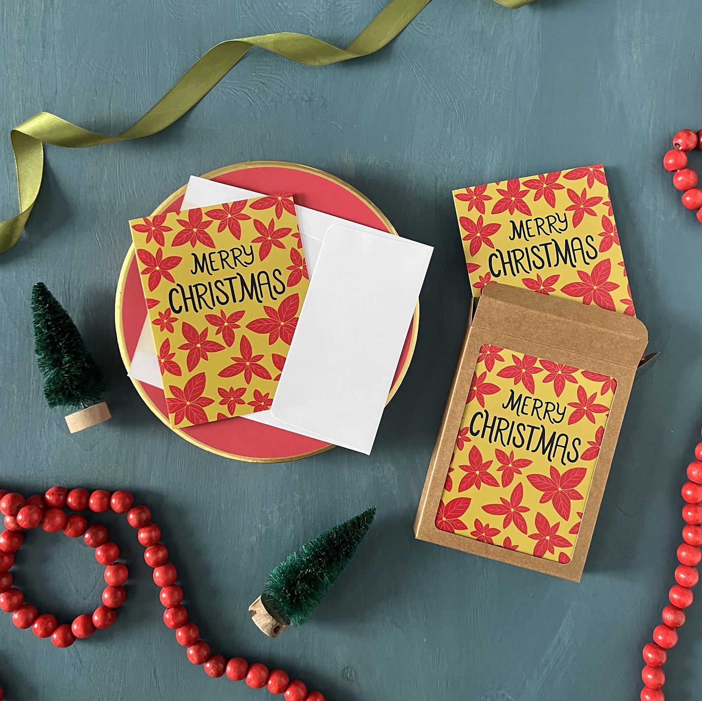 Greeting cards patterned with poinsettias and reading 'Merry Christmas' appear against a festive blue background with green ribbon, red beads, and bottle brush trees.