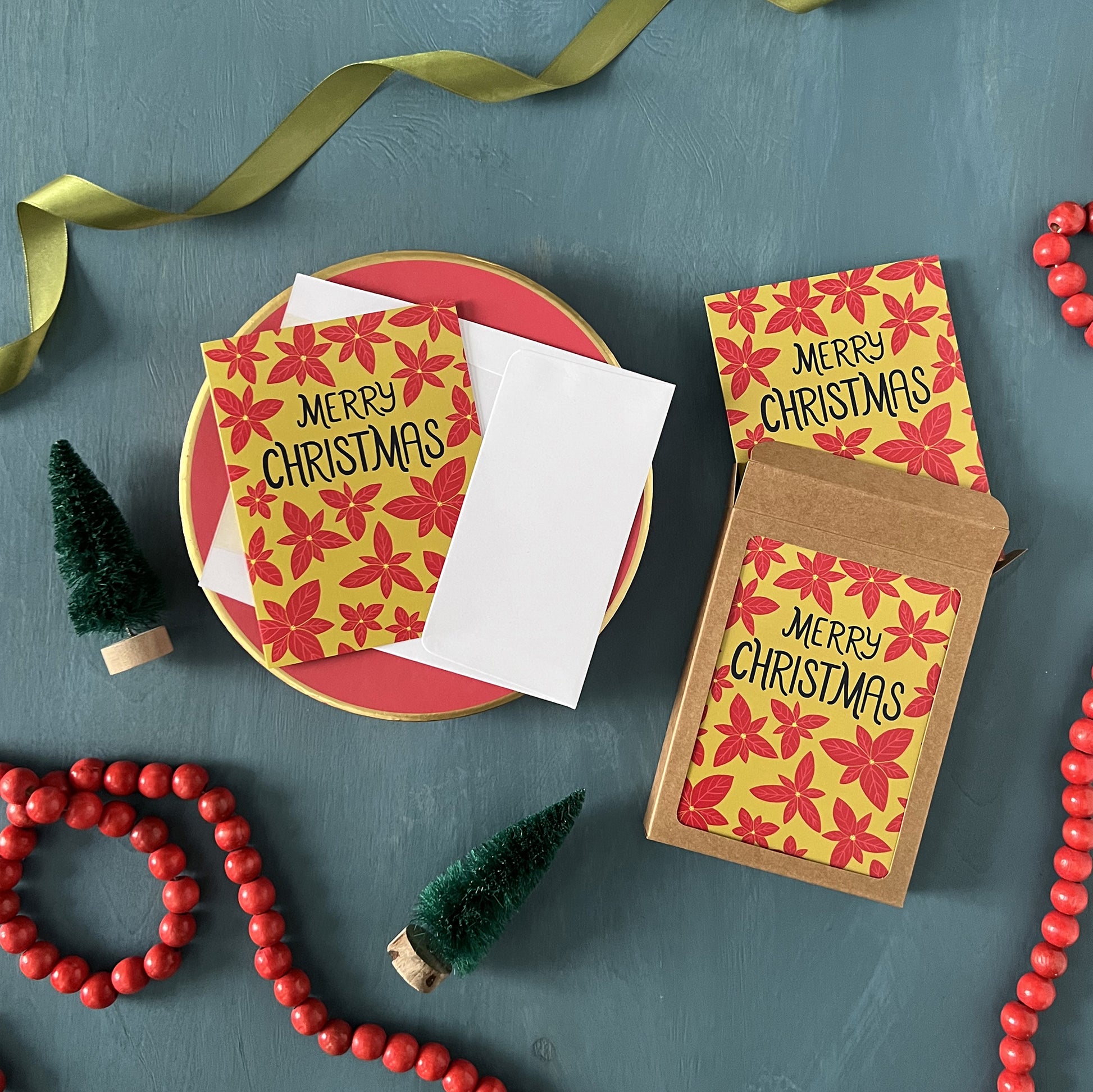 Greeting cards patterned with poinsettias and reading 'Merry Christmas' appear against a festive blue background with green ribbon, red beads, and bottle brush trees.