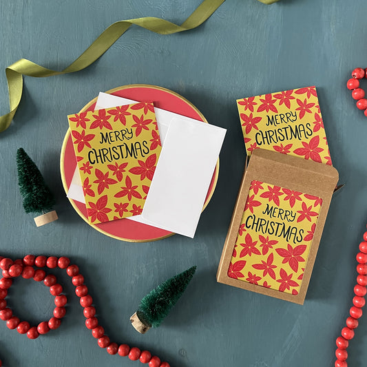 Greeting cards patterned with poinsettias and reading 'Merry Christmas' appear against a festive blue background with green ribbon, red beads, and bottle brush trees.