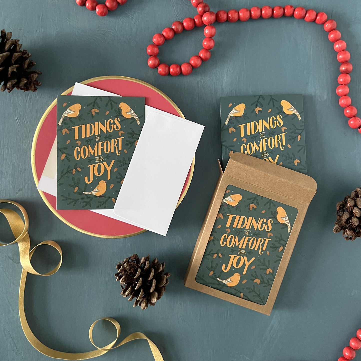 Christmas greeting cards that show goldfinches and the words 'tidings of comfort and joy' appear on a festive blue background with red beads, gold ribbon, and pinecones.