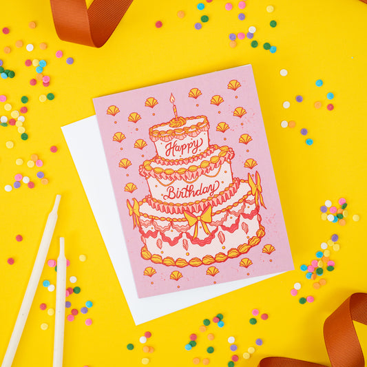 A pink birthday card shows tiered rococo cake and Happy Birthday lettering. It is on a yellow background with confetti and ribbon.