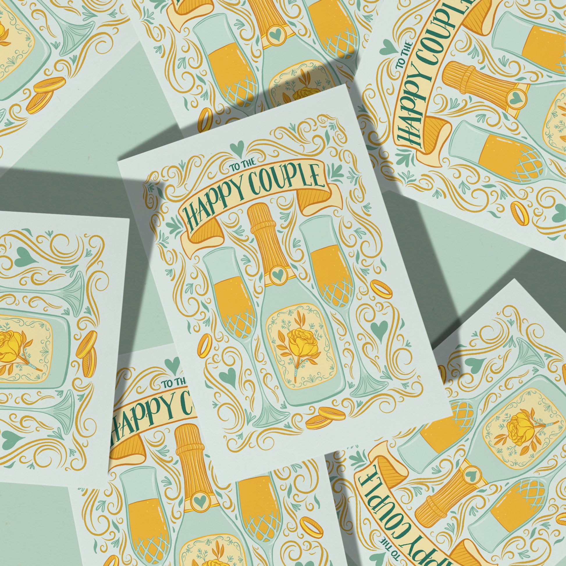  pastel blue greeting card features an illustration of two full champagne flutes, a champagne bottle with a yellow rose, wedding bands, Art Nouveau flourishes, hearts, and the words To The Happy Couple. The card is atop a pile of copies of the same card against a mint background.