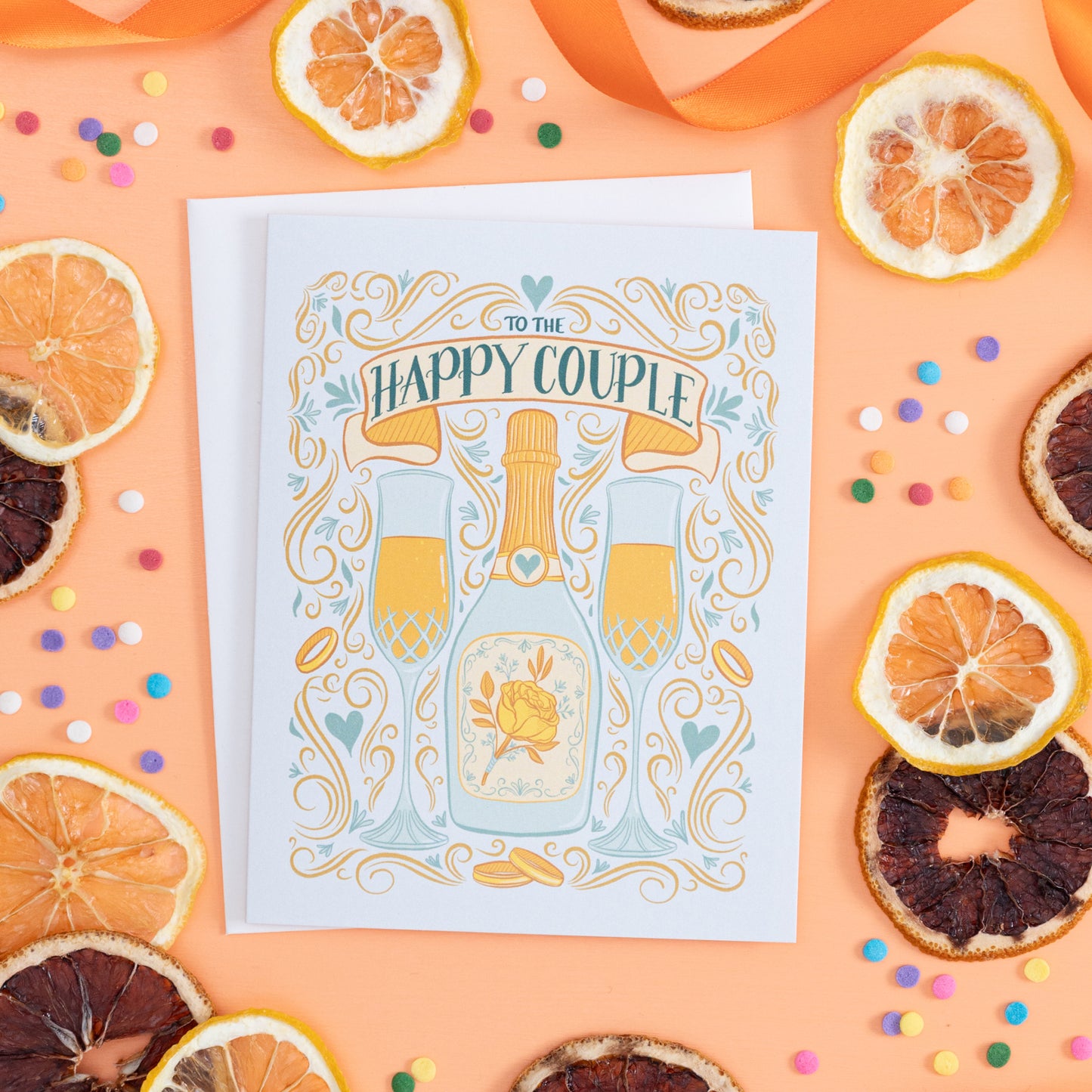 A wedding card with flourishes and hand lettering is on an orange background with citrus slices and confetti.