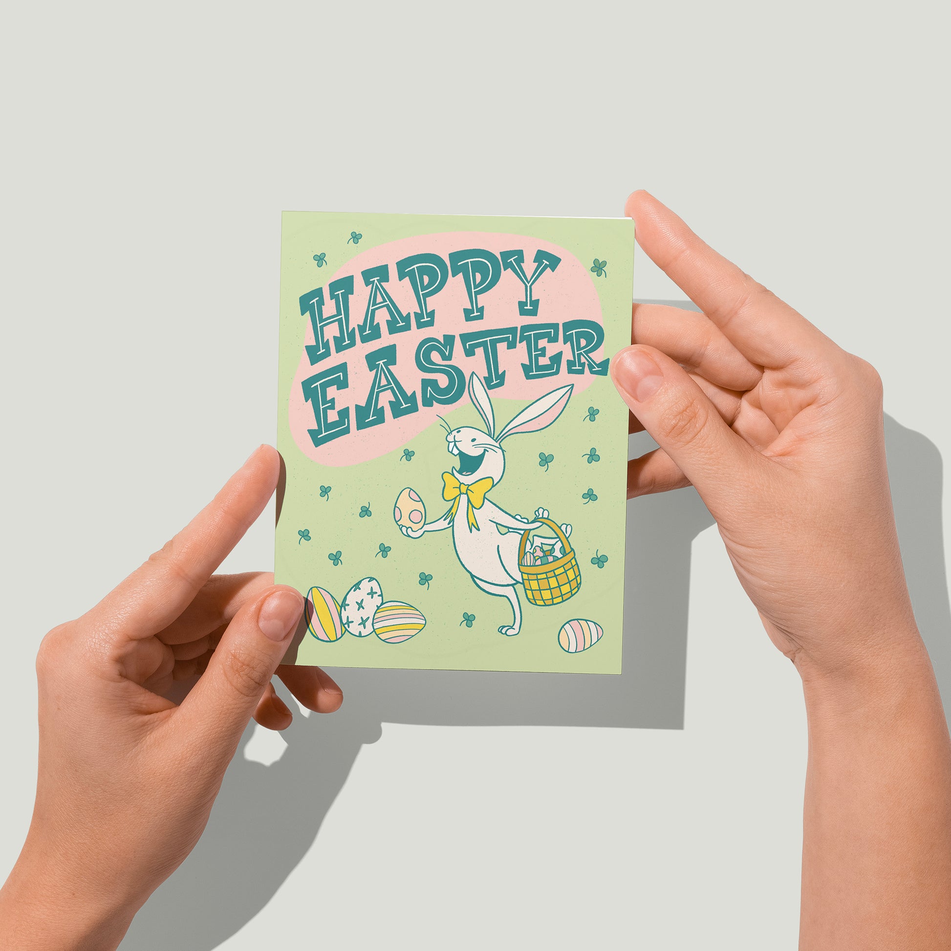 A pale green greeting card shows a cheerful white rabbit wearing a yellow bow tie holding a polka dotted easter egg in one paw and a basket of eggs in the other. There are easter eggs in front of and behind the rabbit. The card reads Happy Easter in midcentury-inspired hand-lettering and shows a shower of green clover. A pair of hands holds the card up in front of the camera.
