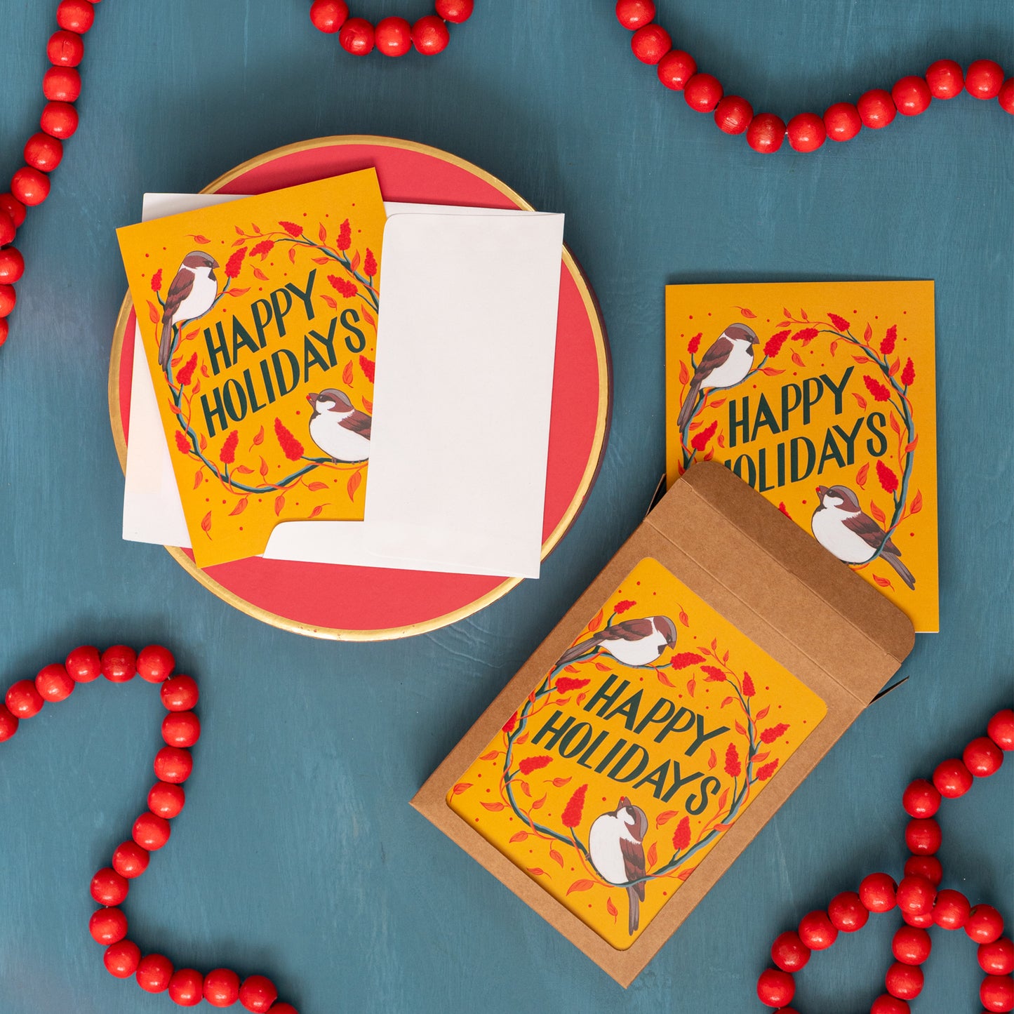 A yellow 'Happy Holidays' card with sparrows and sumac against a festive blue background with red beads and a box of the same Christmas card.