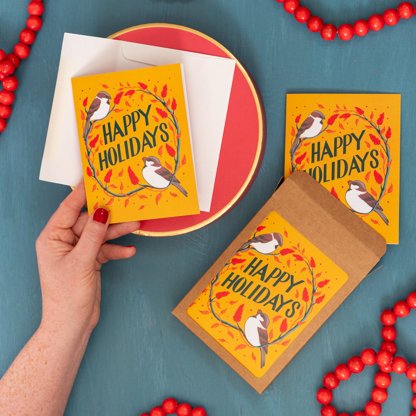 A hand holds a yellow 'Happy Holidays' card with sparrows and sumac against a festive blue background with red beads and a box of the same Christmas card.
