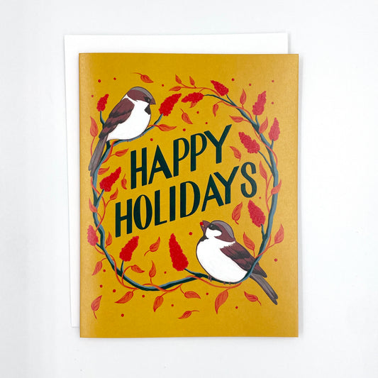 A yellow greeting card features the words Happy Holidays in hand lettering, surrounded by sparrows and staghorn sumac. The card is surrounded by red and gold ornaments, pinecones, and greenery.