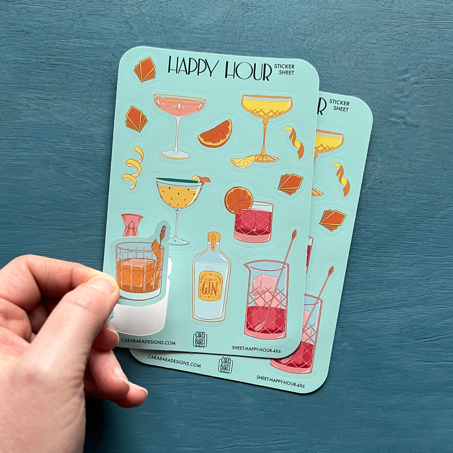 Two art deco cocktail-themed sticker sheets are on a blue background. A hand lifts the old-fashioned sticker off the front sheet.