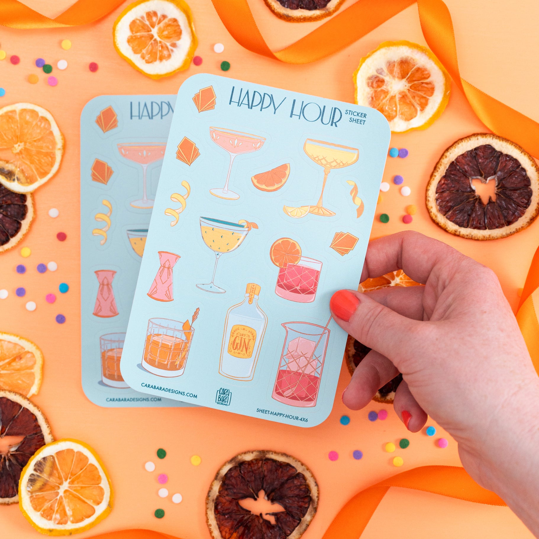 Two art deco cocktail-themed sticker sheets are on an orange background with citrus slices. A hand lifts the top sticker sheet up.