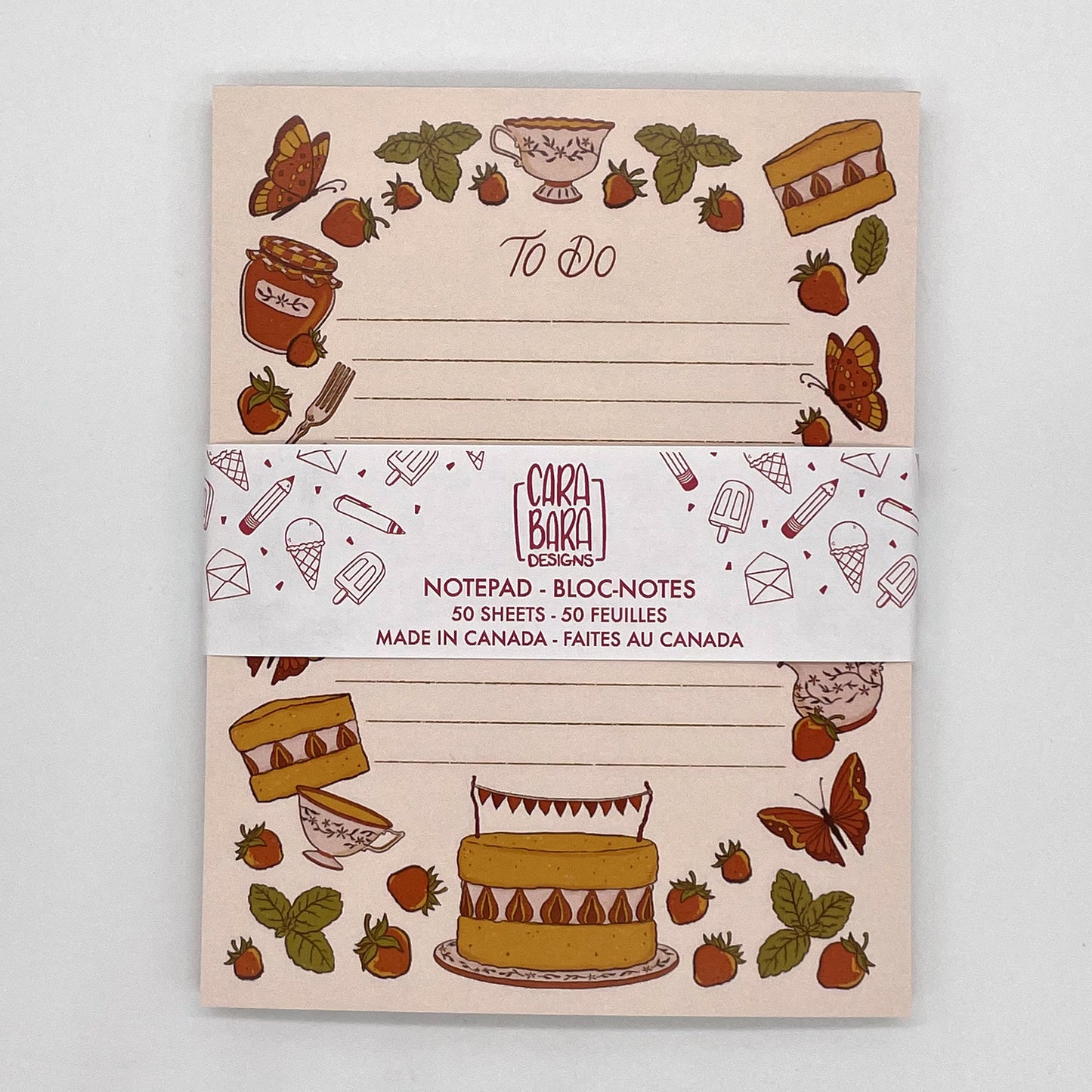 A pink to-do lined cottagecore notepad features illustrated strawberries, mint, cake, tea, and butterflies. It is packaged with a belly band featuring the Carabara Designs logo and indicating that it is 50 pages and made in Canada.