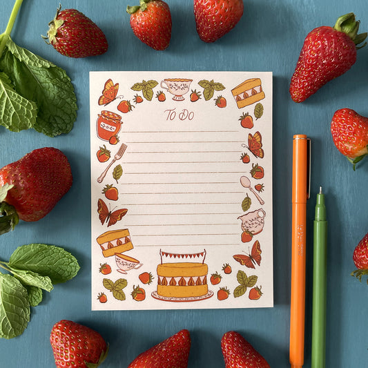 A pink to-do list noepad features illustrated strawberries, mint, cake, tea and butterflies. The 50-page notepad is surrounded by strawberries, mint leaves, and orange and green pens.