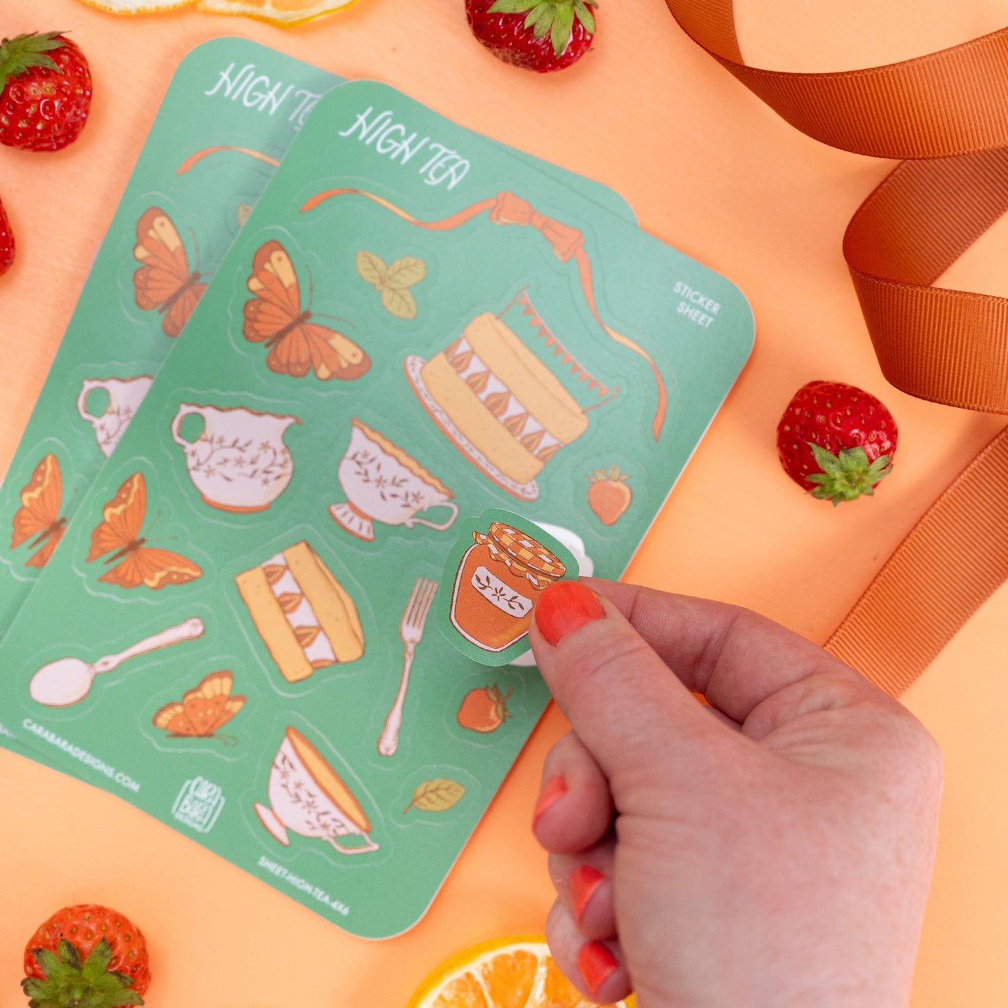 Two cottagecore high tea-themed sticker sheets are on orange with strawberries and ribbon. A hand lifts a jam sticker off the front sheet.
