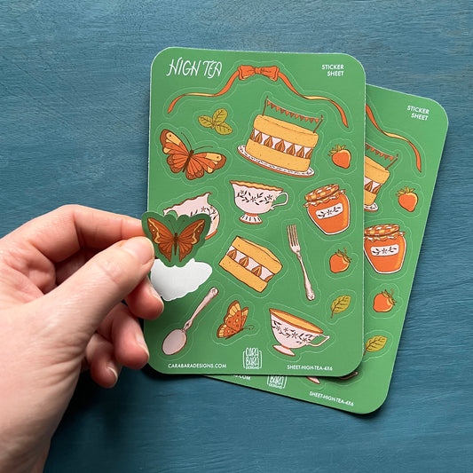 Two cottagecore high tea-themed sticker sheets are on a blue background. A hand lifts a butterfly sticker off the front sheet.