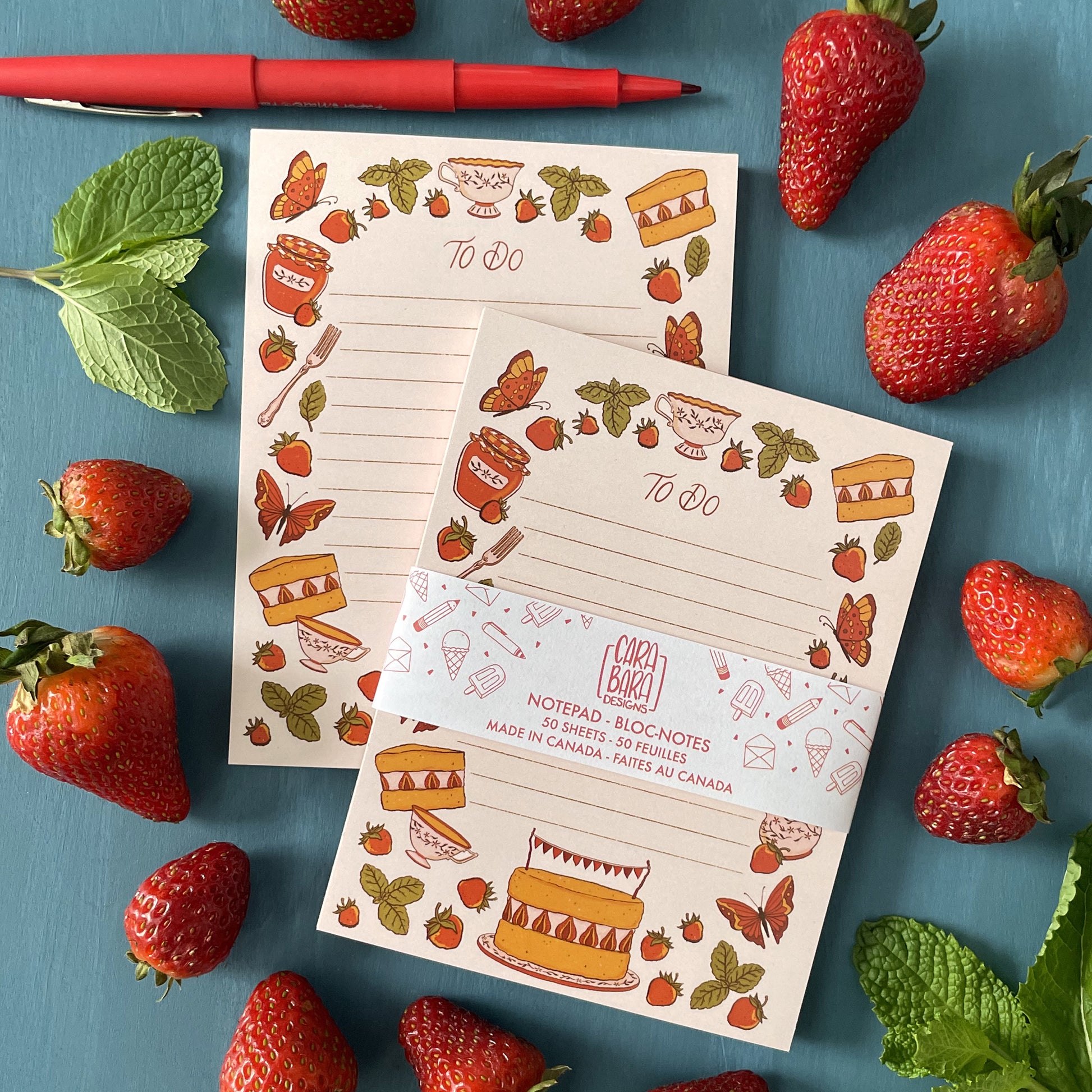 A pair of pink to-do lined cottagecore notepad feature illustrated strawberries, mint, cake, tea, and butterflies. The front notepad is packaged with a belly band featuring the Carabara Designs logo and indicating that it is 50 pages and made in Canada. The notepads are surrounded by strawberries, mint leaves, and a red pen.