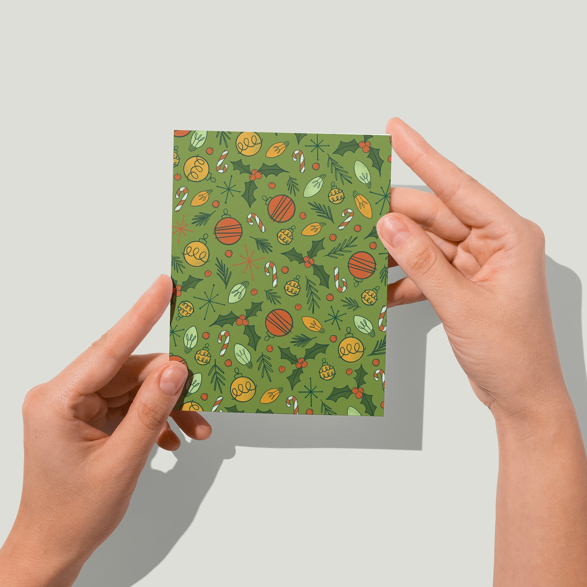 A green greeting card is patterned with midcentury-inspired ornaments, holly, candy canes and Christmas lights. A pair of hands holds the card up in front of the camera.