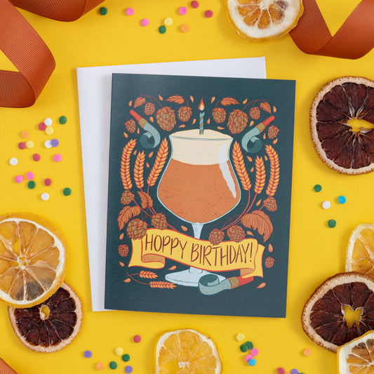 A beer-themed birthday card with the words 'hoppy birthday' is on yellow with confetti and citrus slices.