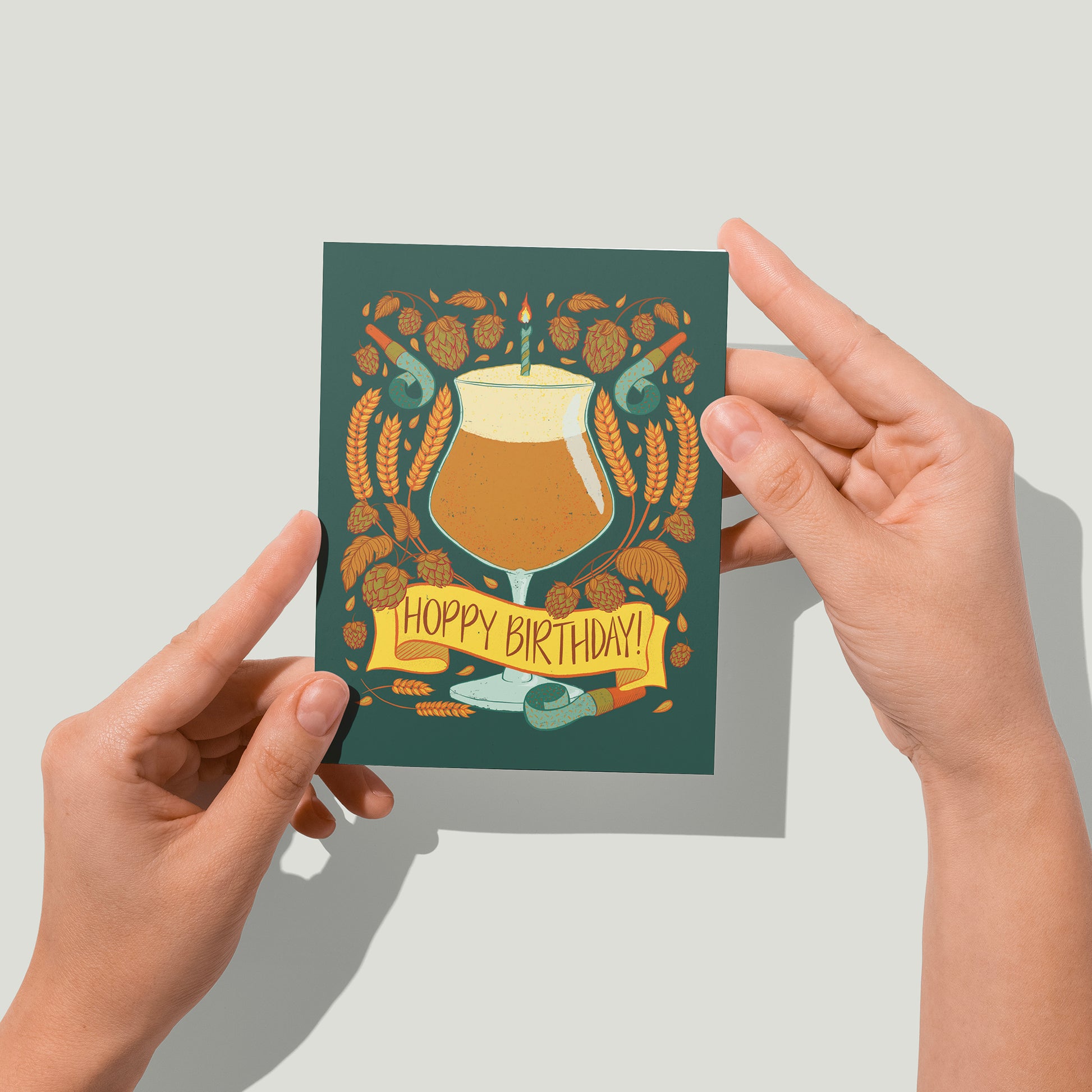 A dark green greeting card features an illustration of a full and frothy beer glass with a birthday candle in it. At the bottom of the glass, a yellow banner reads Hoppy Birthday. Around the glass are stalks of wheat, wheat berries, hops blossoms, and light blue noisemakers. A pair of hands holds the card up in front of the camera.