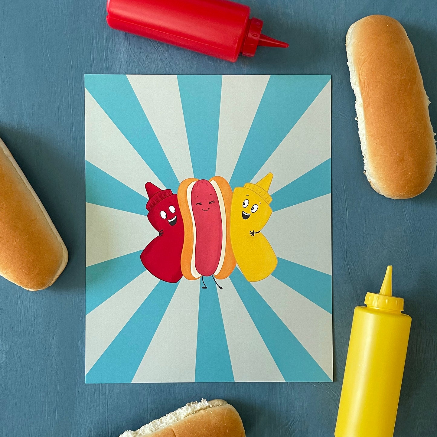 An art print shows cartoon ketchup, mustard, and hot dog in a bun on a blue background. The print is on a blue background with hot dog buns and squeeze bottles.