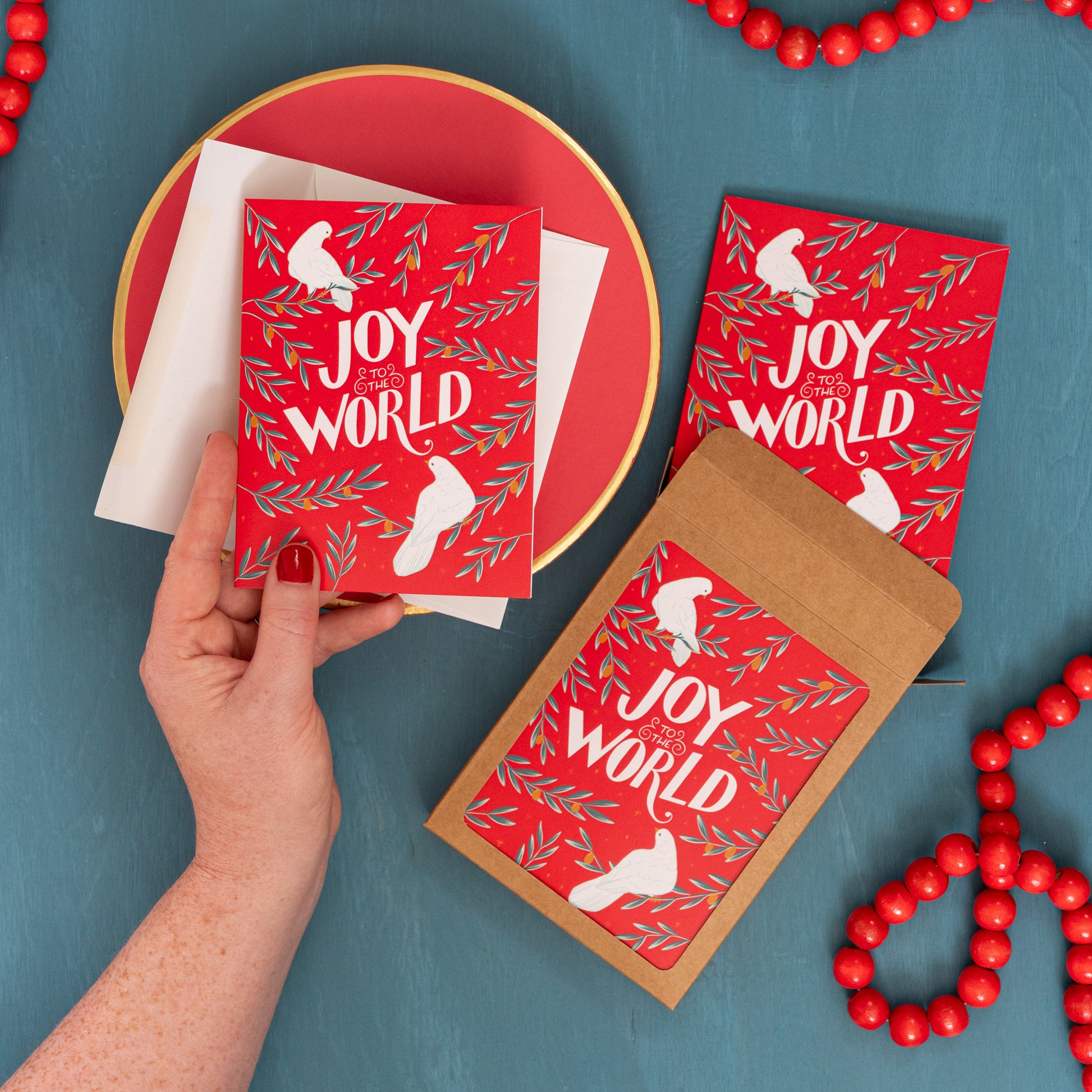 A hand holds a red 'Joy to the World' card with doves and olive branches against a festive blue background with red beads and a box of the same Christmas card.