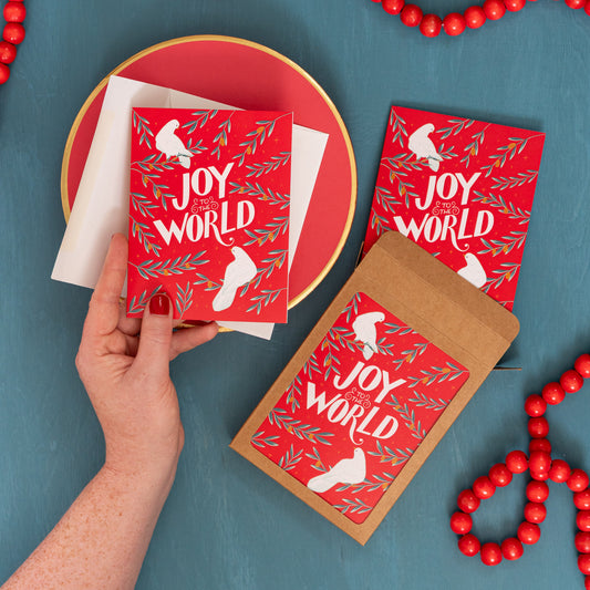 A hand holds a red 'Joy to the World' card with doves and olive branches against a festive blue background with red beads and a box of the same Christmas card.