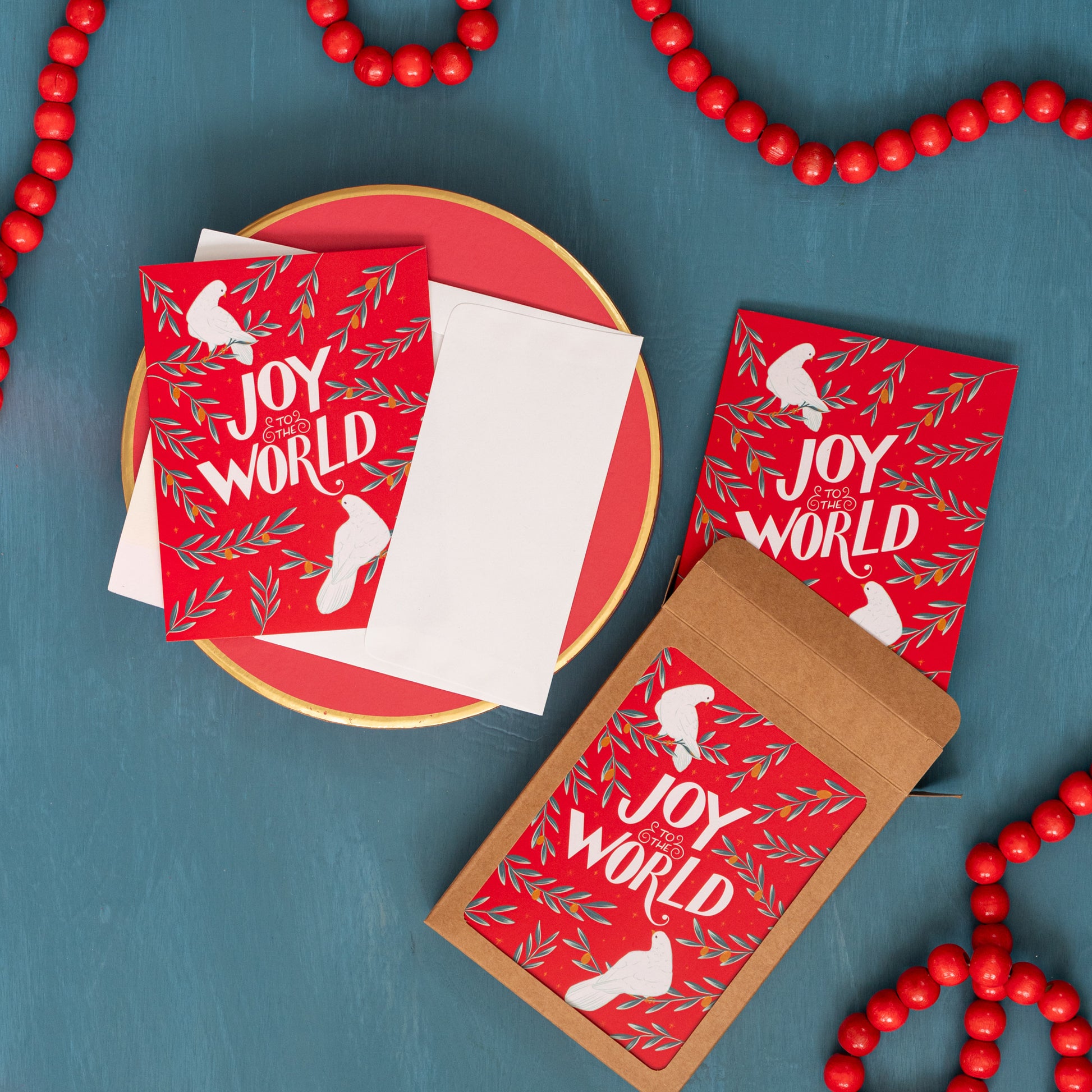 A red 'Joy to the World' card with doves and olive branches appears against a festive blue background with red beads and a box of the same Christmas card.