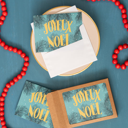 A blue Christmas card showing the words 'Joyeux Noel' appears against a festive blue background with red beads and a box of the same card.