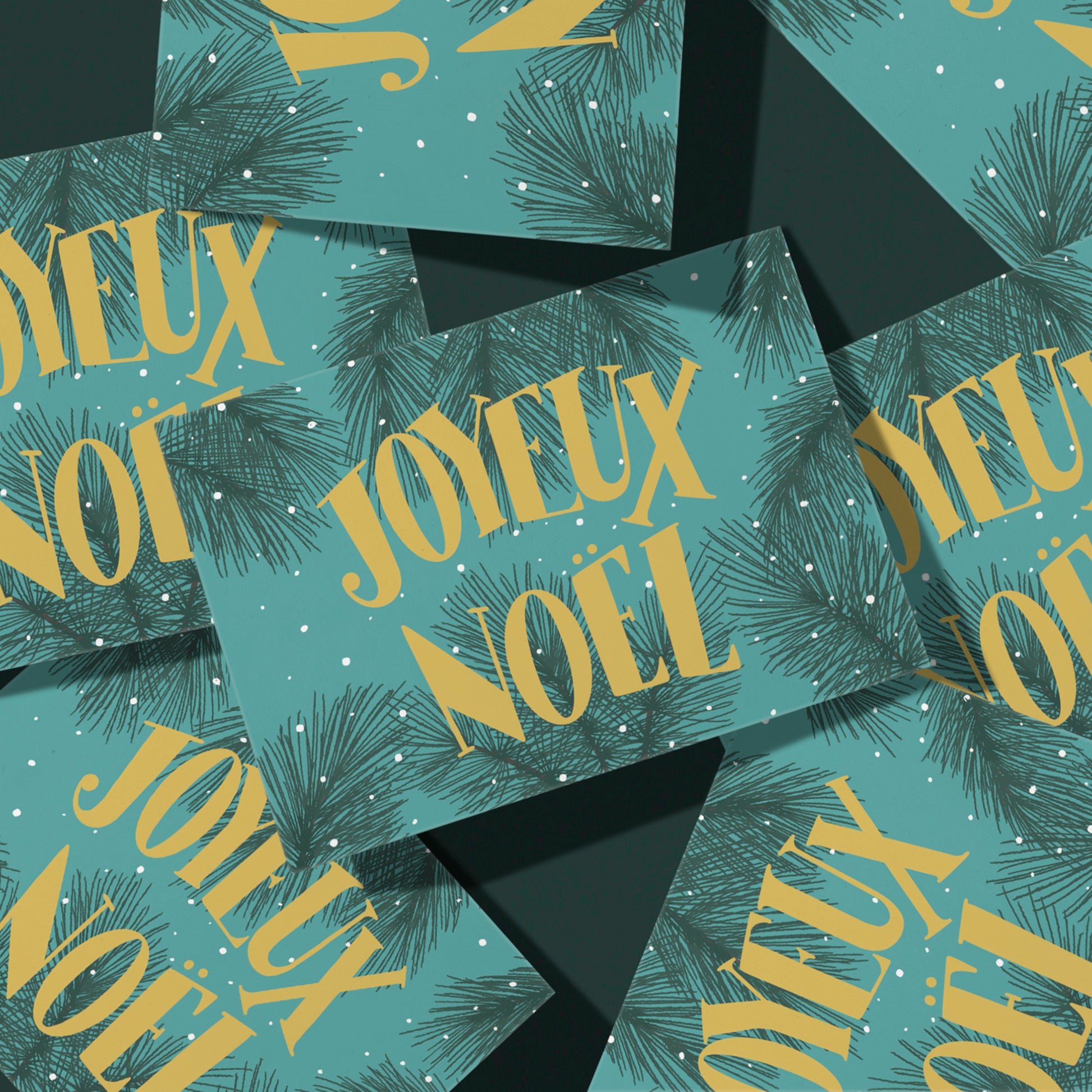 A teal greeting card features the words Joyeux Noel in midcentury-inspired yellow lettering against a background of snowy evergreen branches. The card sits atop a pile of copies of the same card on a dark green background.